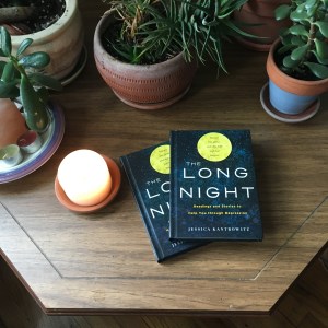 Two copies of The Long Night lying on a table with plants and candles